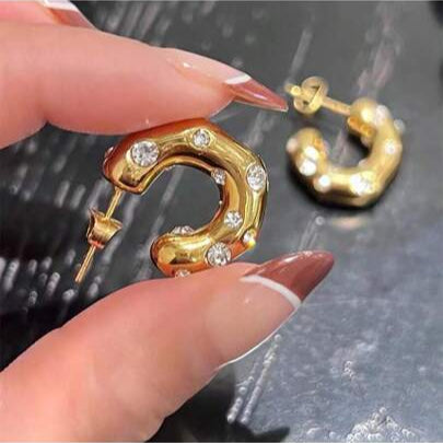 Gold earrings with clear stones held by a hand against a dark background