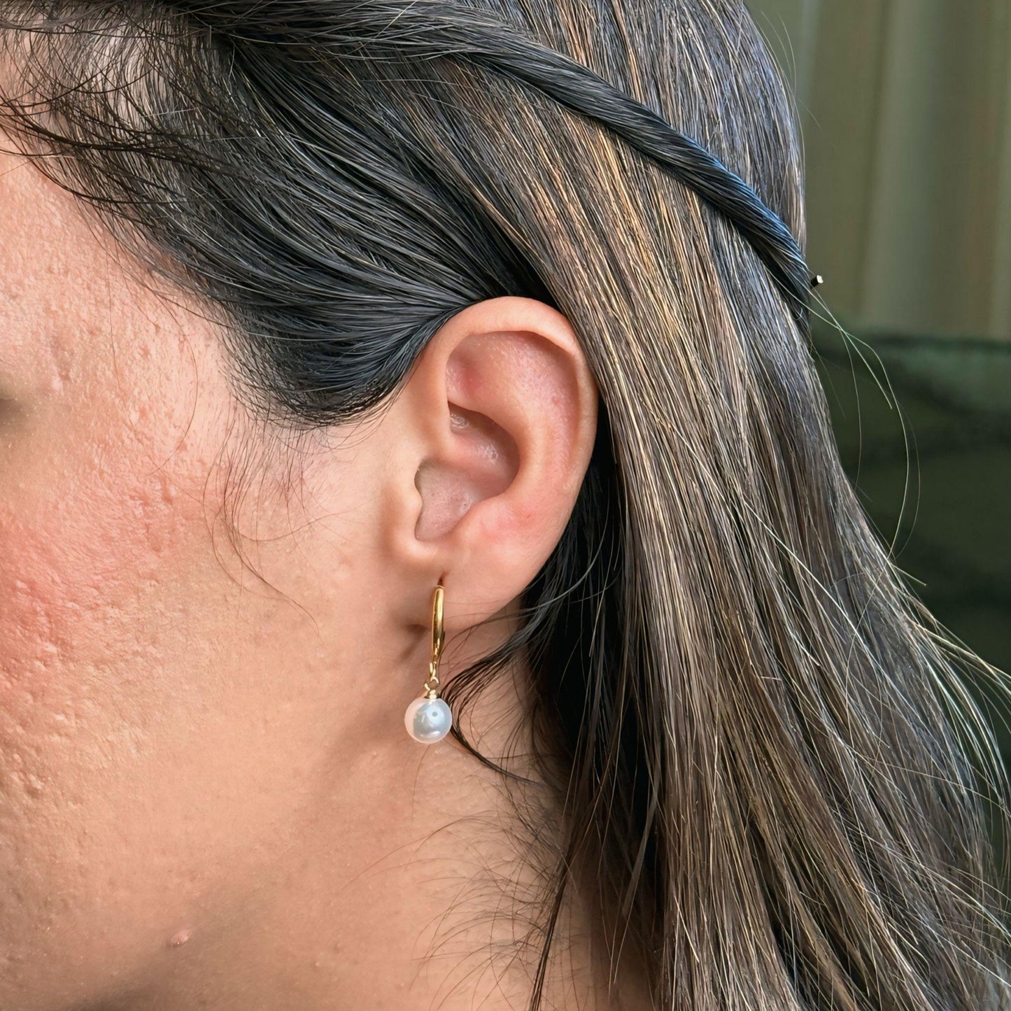 Close-up of a person wearing a gold earring with a pearl, against a neutral background.